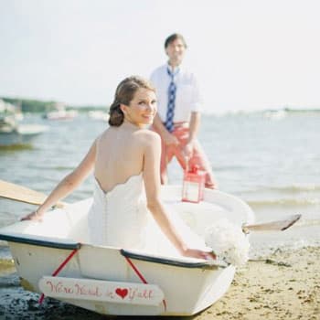 Невеста и жених на свадебной фотосессии у берега в лодке с табличкой We're hitched y'all