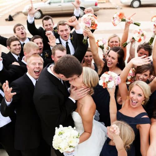 Жених и невеста целуются на фоне радостных гостей, которые поднимают руки и букеты в воздух, создавая атмосферу праздника