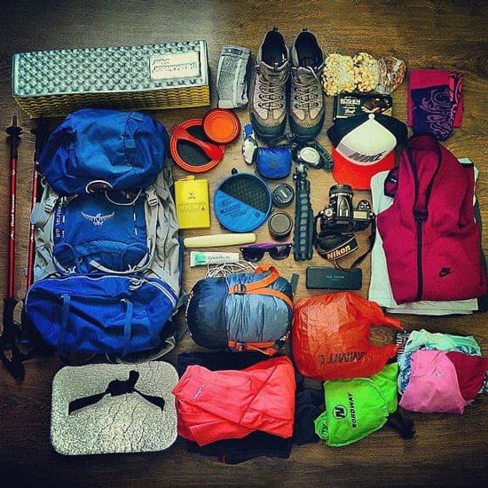 Разложенные на полу туристические вещи, включая рюкзаки, обувь, одежду, снаряжение и фотокамеру, подготовленные для похода