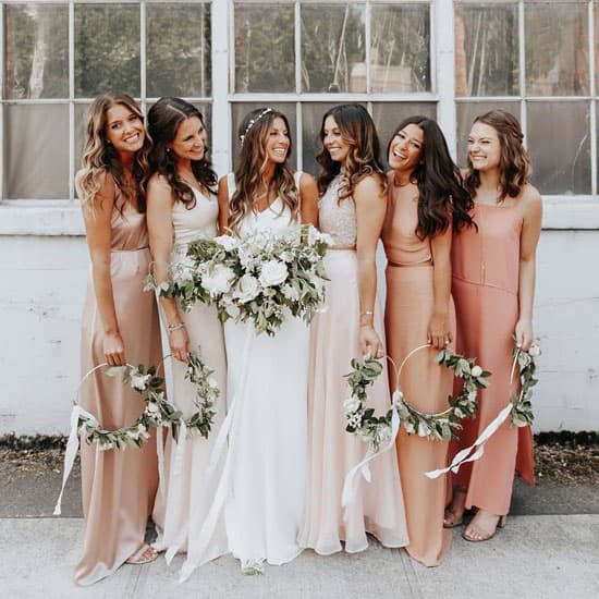 Невеста в белом платье с букетом и подружки невесты в нежных платьях пастельных тонов позируют вместе, улыбаются и держат венки из зелени