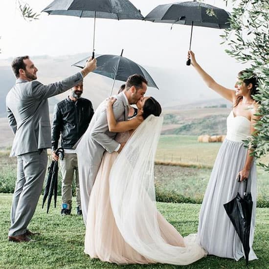 Жених и невеста целуются под дождём на фоне живописного пейзажа, а друзья держат над ними чёрные зонты